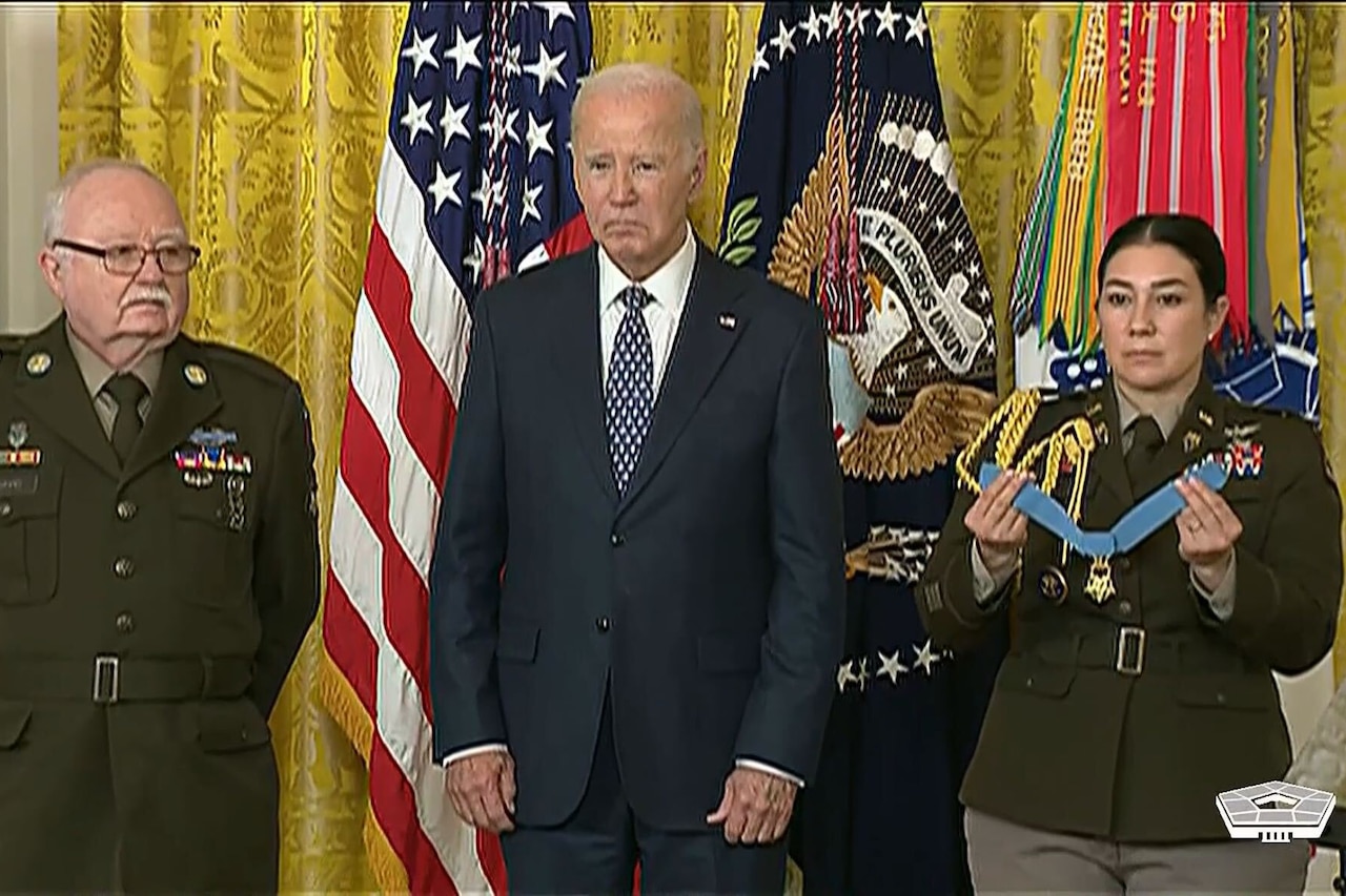 Three people stand on a stage in front of flags.