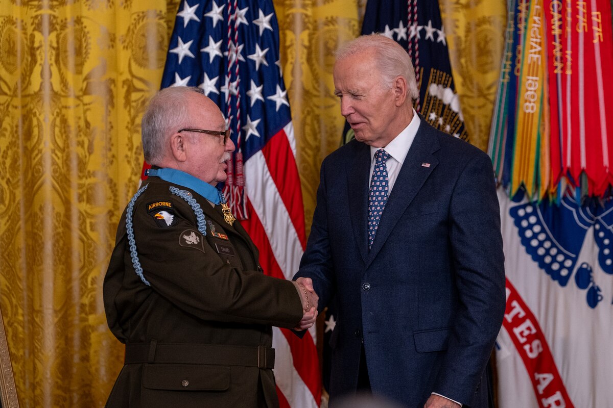 President Joe Biden shakes hands with a veteran soldier, with flags in the background.