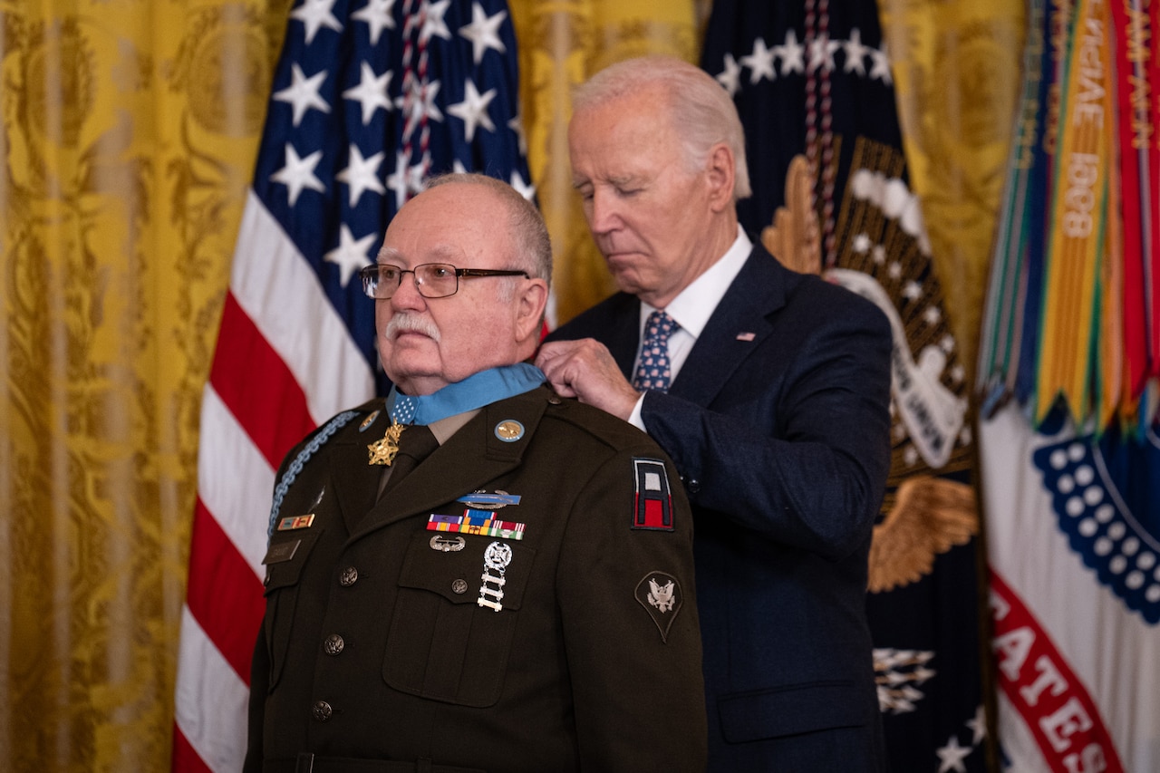 President Joe Biden places the Medal of Honor around the neck of a veteran soldier, with flags in the background.