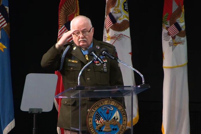 Army Pfc. Kenneth J. David wears the Medal of Honor while saluting from a lectern with flags on a black background.