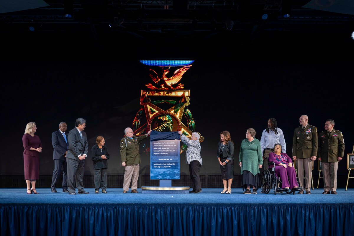 A Medal of Honor recipient and a civilian lift the cloth to unveil a plaque while flanked by several others on a stage.