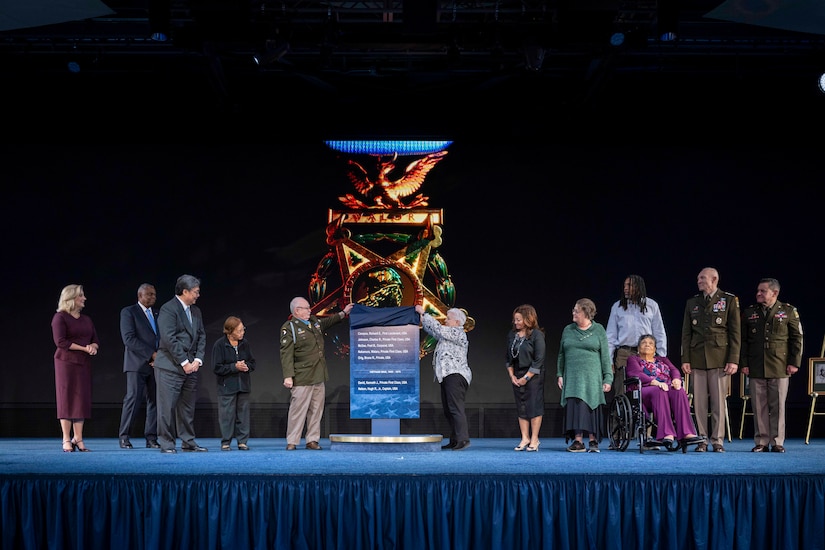 A Medal of Honor recipient and a civilian lift the cloth to unveil a plaque while flanked by several others on a stage.