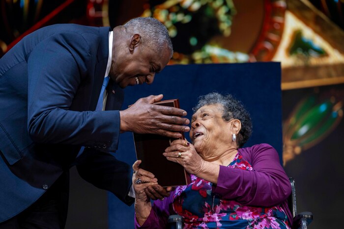 A man in a blue suit presents a woman in a purple dress sitting in a wheelchair a display case.