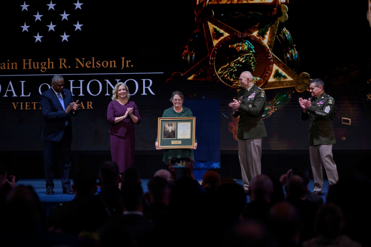 A civilian holding a plaque is flanked by military and defense officials on a stage.