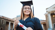 A young black woman with glasses standing in her cap and gown holding a diploma in front of a college building