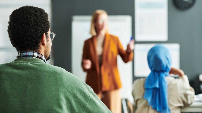 A woman instructor standing in front of a room with a man and woman siting and facing the instructor