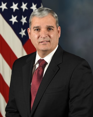 Mr. Bracero poses for his official portrait in front of a blue background with an American flag behind him on the left.