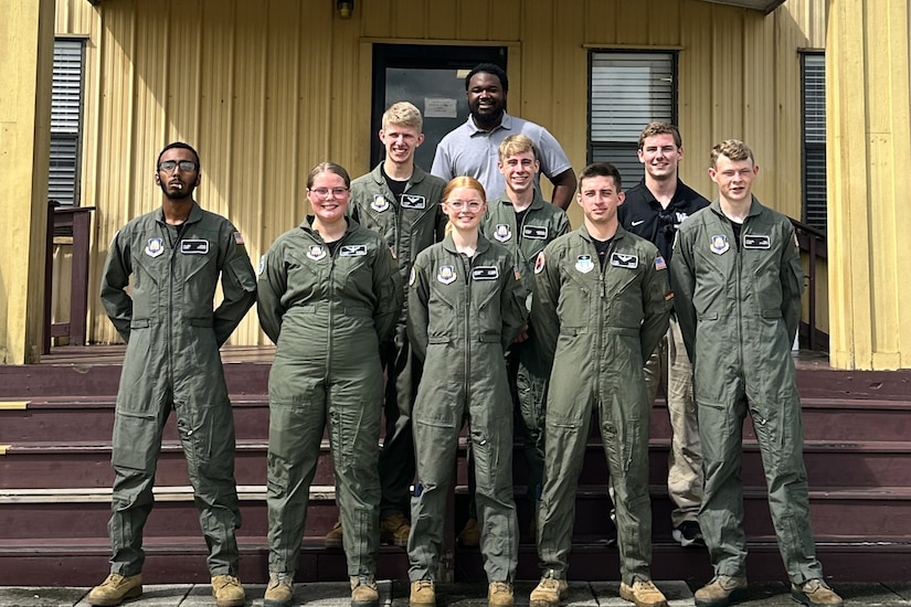 Seven cadets and two instructors smile in front of a yellow building.