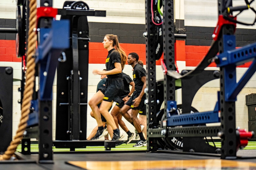 A group of soldiers run on a track inside a gym with weightlifting equipment in the foreground.