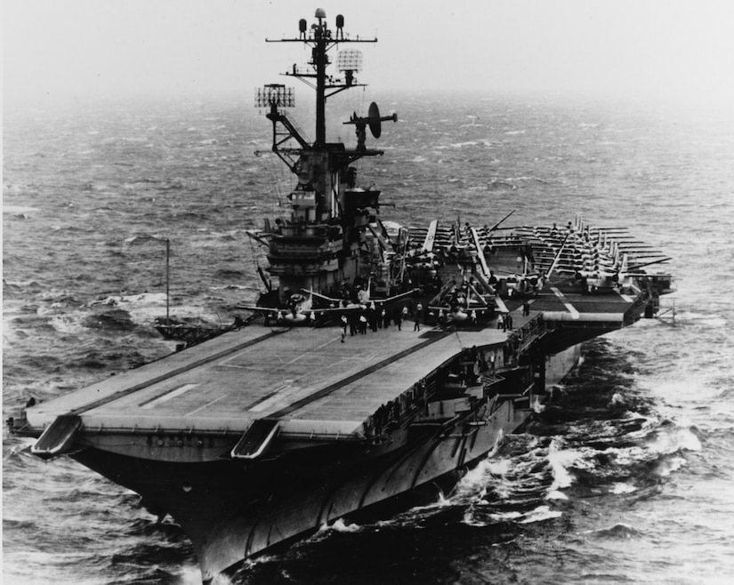 In black and white, several small aircraft sit on the deck of a large ship with sailors on deck.