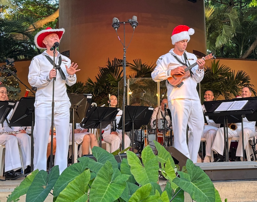 A sailor sings into a microphone on stage with a band while another plays the ukulele.