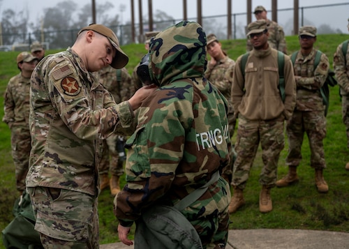 U.S. Air Force Tech. Sgt. Zachary Hancock, 311th Training Squadron instructor, assists an Airmen don on protective gear during chemical, biological, radiological, and nuclear response training during a deployment readiness exercise, Presidio of Monterey, Calif., Jan. 6, 2025. Mission-Oriented Protective Posture gear is designed to shield service members from harmful agents that could cause severe injury or death. (U.S. Air Force photo by Senior Airman Sarah Williams)