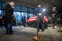 A man bows his head (left) while standing near the flag-draped casket of former President Jimmy Carter while it is lying in repose and being guarded by a team of ceremonial guard members, one from each branch of the armed forces.