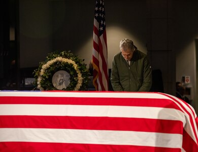 A man bows his head while standing next to a flag-draped casket.