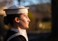 A Navy sailor wearing a white cap is standing at attention during a ceremony.
