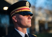 An Army soldier wearing a service hat with gold and red band is participating in a ceremony.