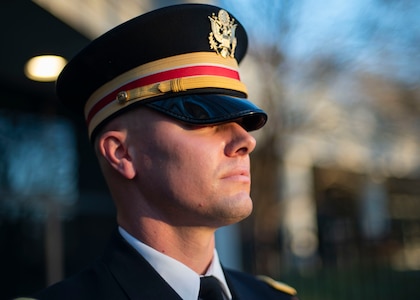 An Army soldier wearing a service hat with gold and red band is participating in a ceremony.