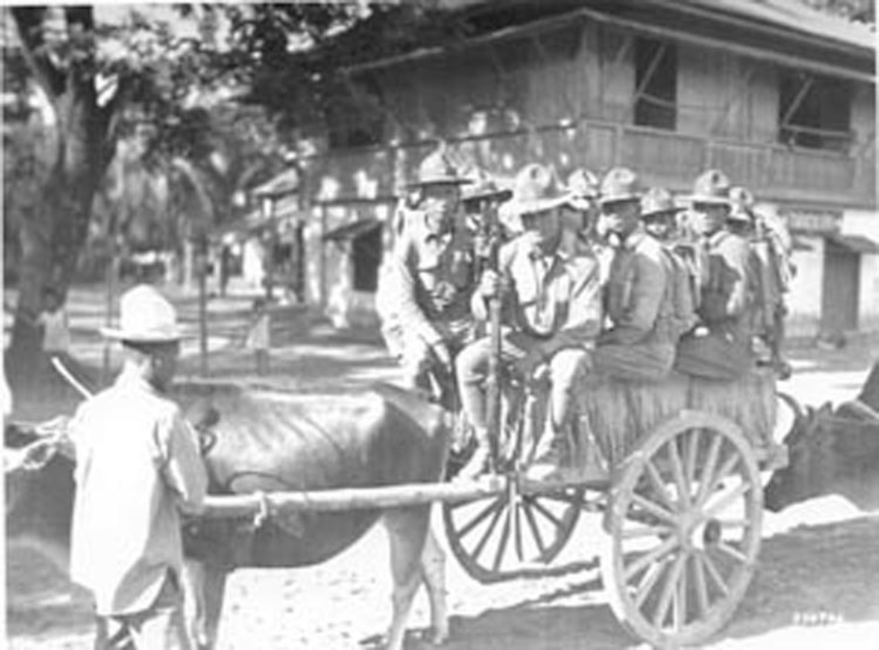 Eight men sit on a small cart attached to cattle.