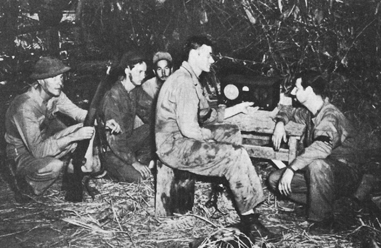 Five men sit in a hay-covered area listening to a radio.