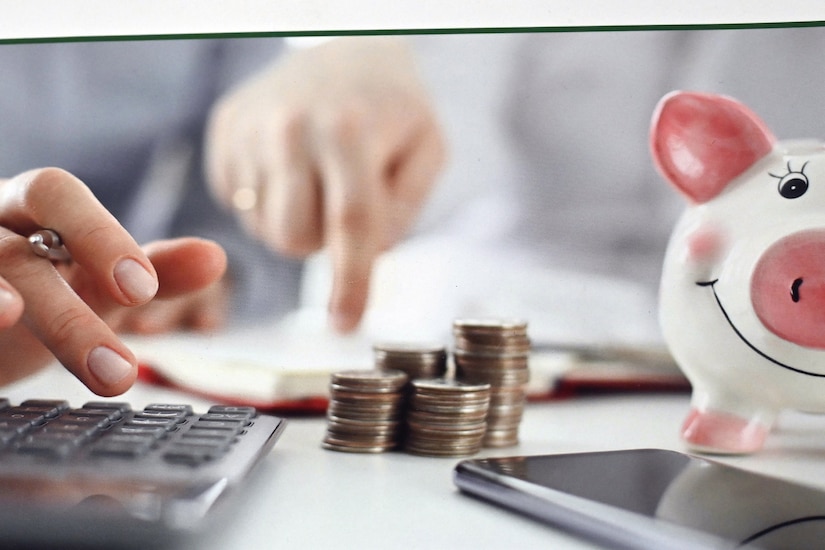 One hand points at a desk while the other types on a calculator near a pile of coins and a piggy bank.