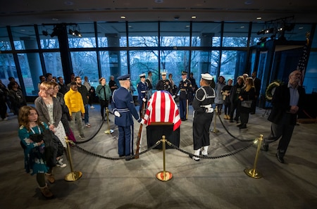 Mourners stand with military servicemembers around a flag-draped casket.