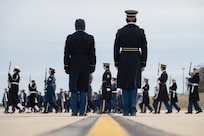 Two service members stand in formation while watching dozens of others march while carrying ceremonial rifles on a gloomy day.