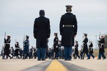 Two service members stand in formation while watching dozens of others march while carrying ceremonial rifles on a gloomy day.