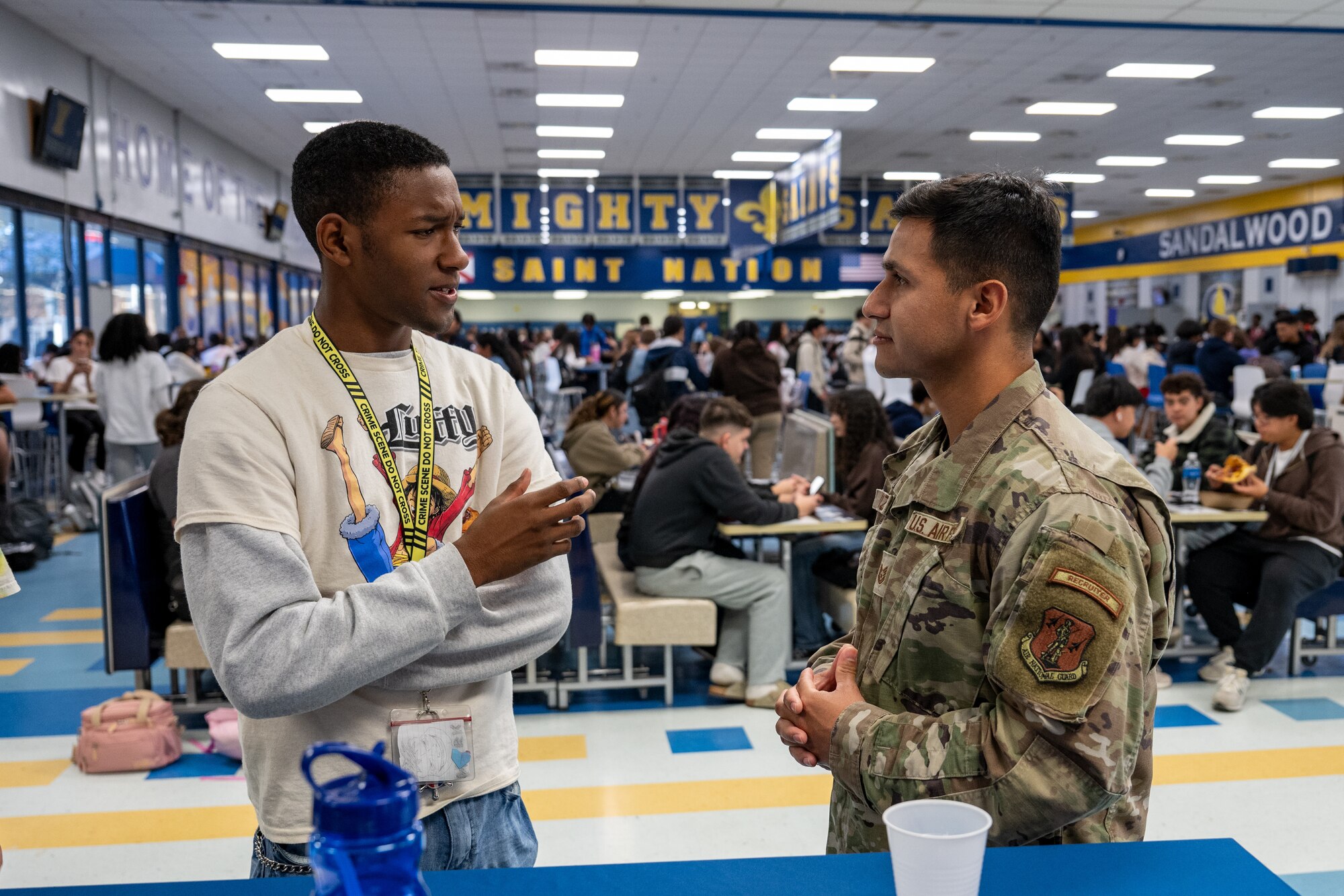A student in a white and gray long-sleeved shirt speaks to a U.S. Air National Guard recruiter in a high school cafeteria.
