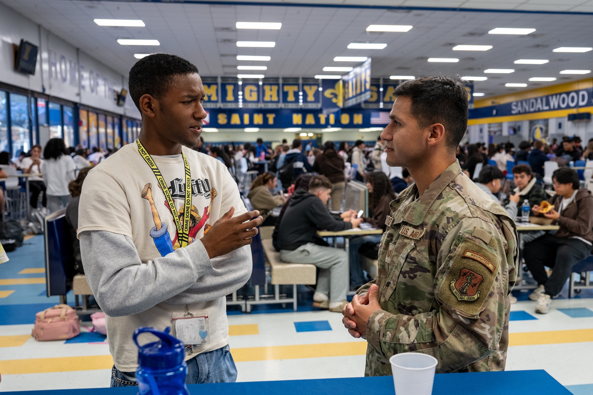 A student in a white and gray long-sleeved shirt speaks to a U.S. Air National Guard recruiter in a high school cafeteria.