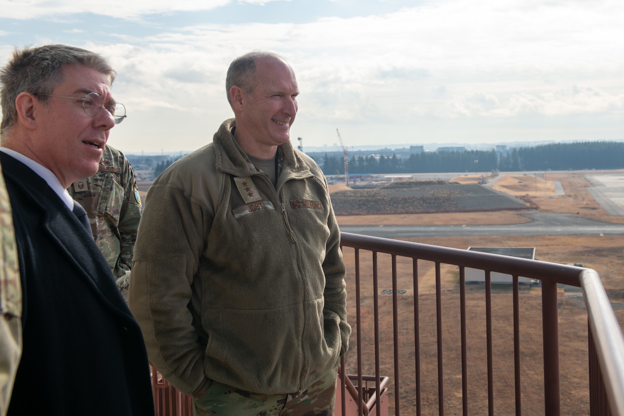 Air Force personnel stand on the catwalk outside of the air traffic control tower and observe the flightline on a sunny dya.