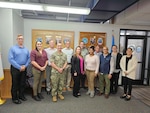 A  large group of people, both men and women stand in a office lobby. One man in front is wearing a Navy military uniform.