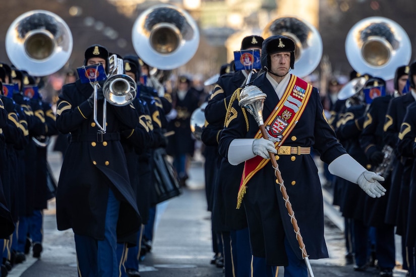 Soldiers playing instruments and wearing winter dress uniforms march in formation down a city street.