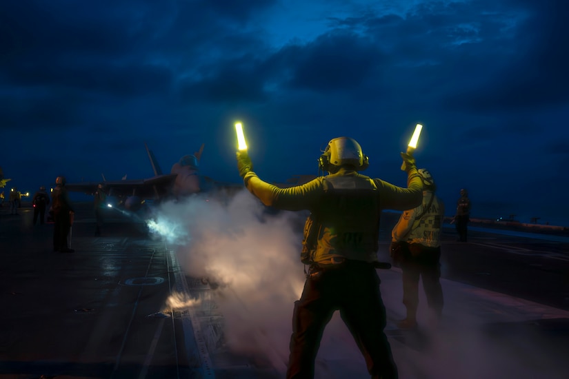 A sailor surrounded by smoke holds two batons illuminated by yellow lights while signaling to an aircraft aboard a ship at sea in the dark.