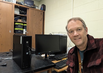A white male looks into the camera. he is wearing red and black plaid shirt. behind him are computers, desktops, screens, and monitors.