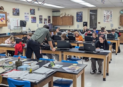 High school students sit at long lab tables working on laptops while a teacher stands looking at them in a class room.