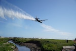 A military airplane is flying over a lush field will white fire suppressant discharging from the back.