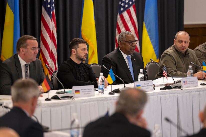 Several men sit at a long table. Tiny flags of various nations adorn the table.