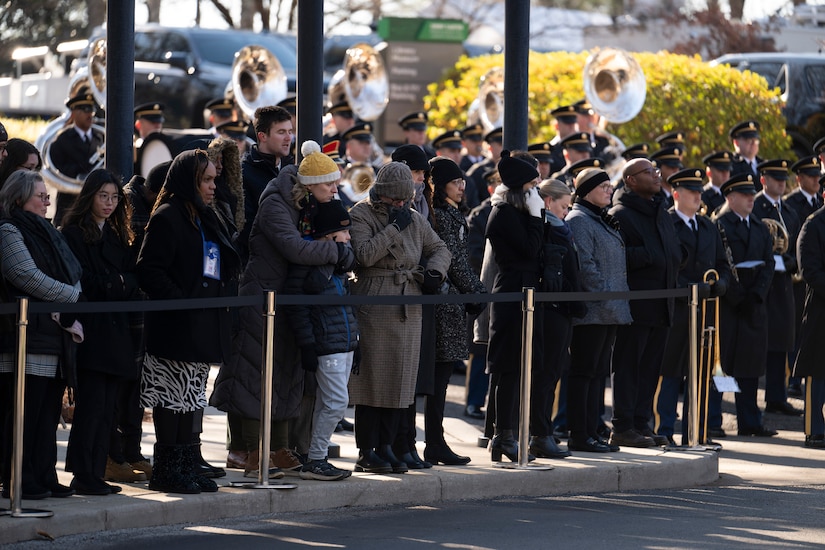 Uniformed service members and civilians stand shoulder-to-shoulder on a sidewalk behind a barrier.