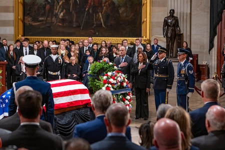 Uniformed service members and civilians in business attire stand in front of a wreath and a flag-draped casket while a large crowd looks on.