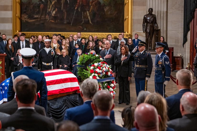 Uniformed service members and civilians in business attire stand in front of a wreath and a flag-draped casket while a large crowd looks on.