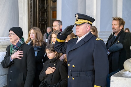 A uniformed service member salutes while several civilians place their right hand over their hearts.