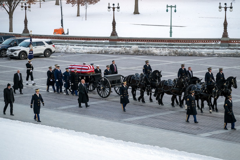 Army soldiers on horseback, wearing winter dress uniforms, pull a caisson loaded with a flag-draped casket down a city street.