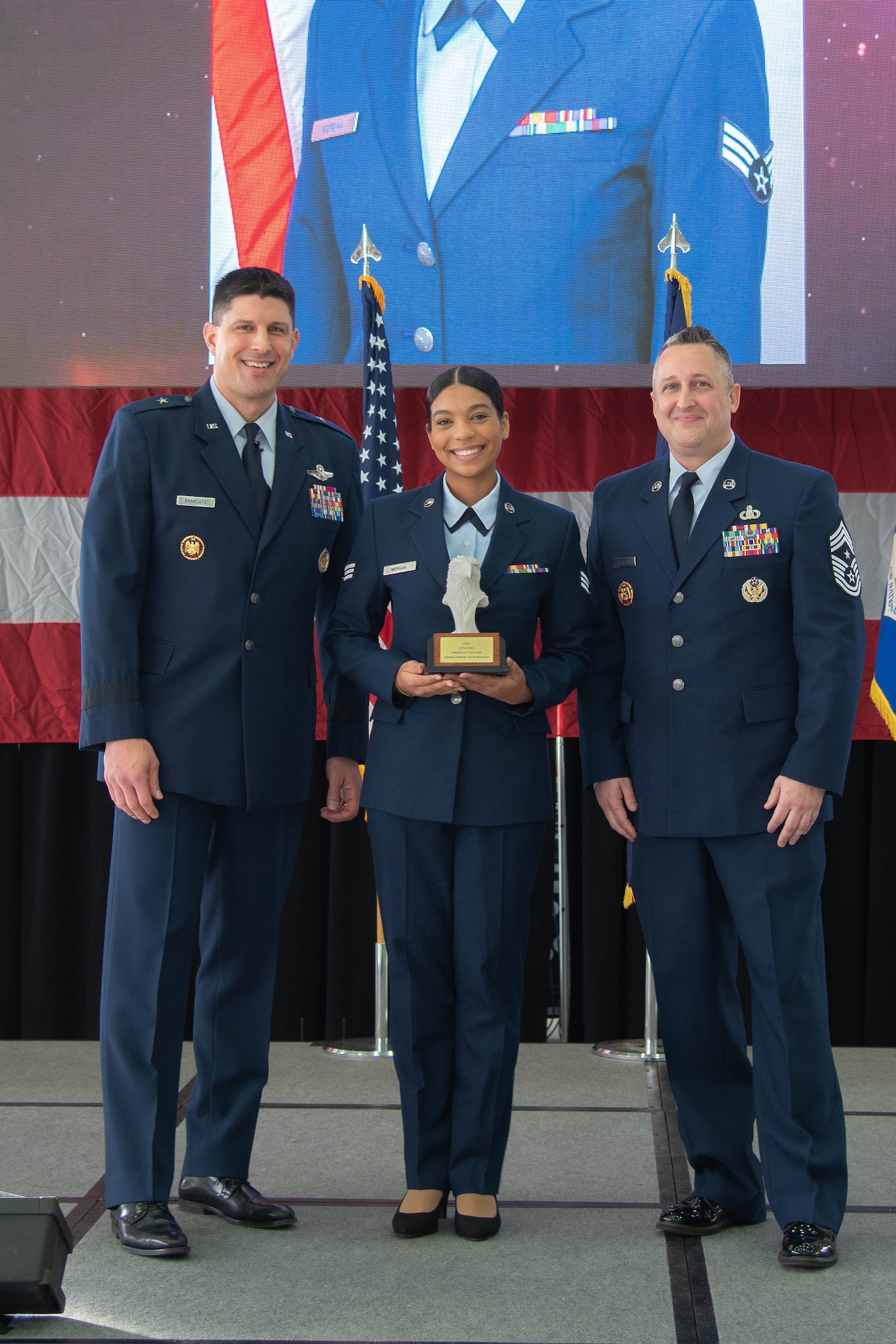 Male Air Force officer in dress blue uniform stands left of Air Force junior enlisted female Airman in dress blue uniform holding trophy, who stands left of Male Air Force senior enlisted leader in dress blue uniform.