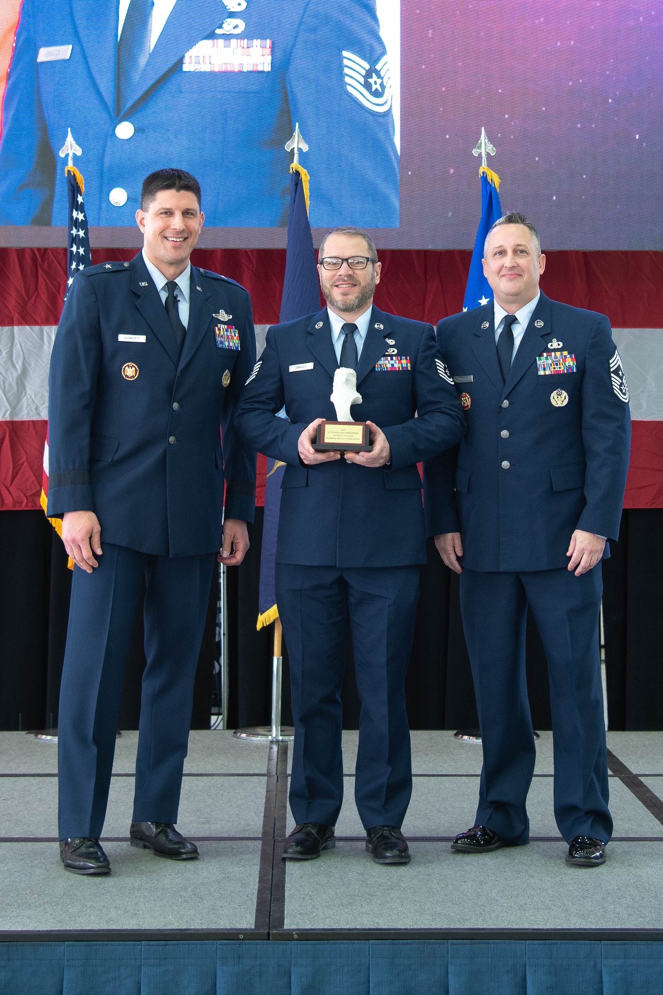 Air Force officer in dress blue uniform stands left of Air Force non-commissioned officer in dress blue uniform holding trophy, who stands left of Air Force senior enlisted leader in dress blue uniform.