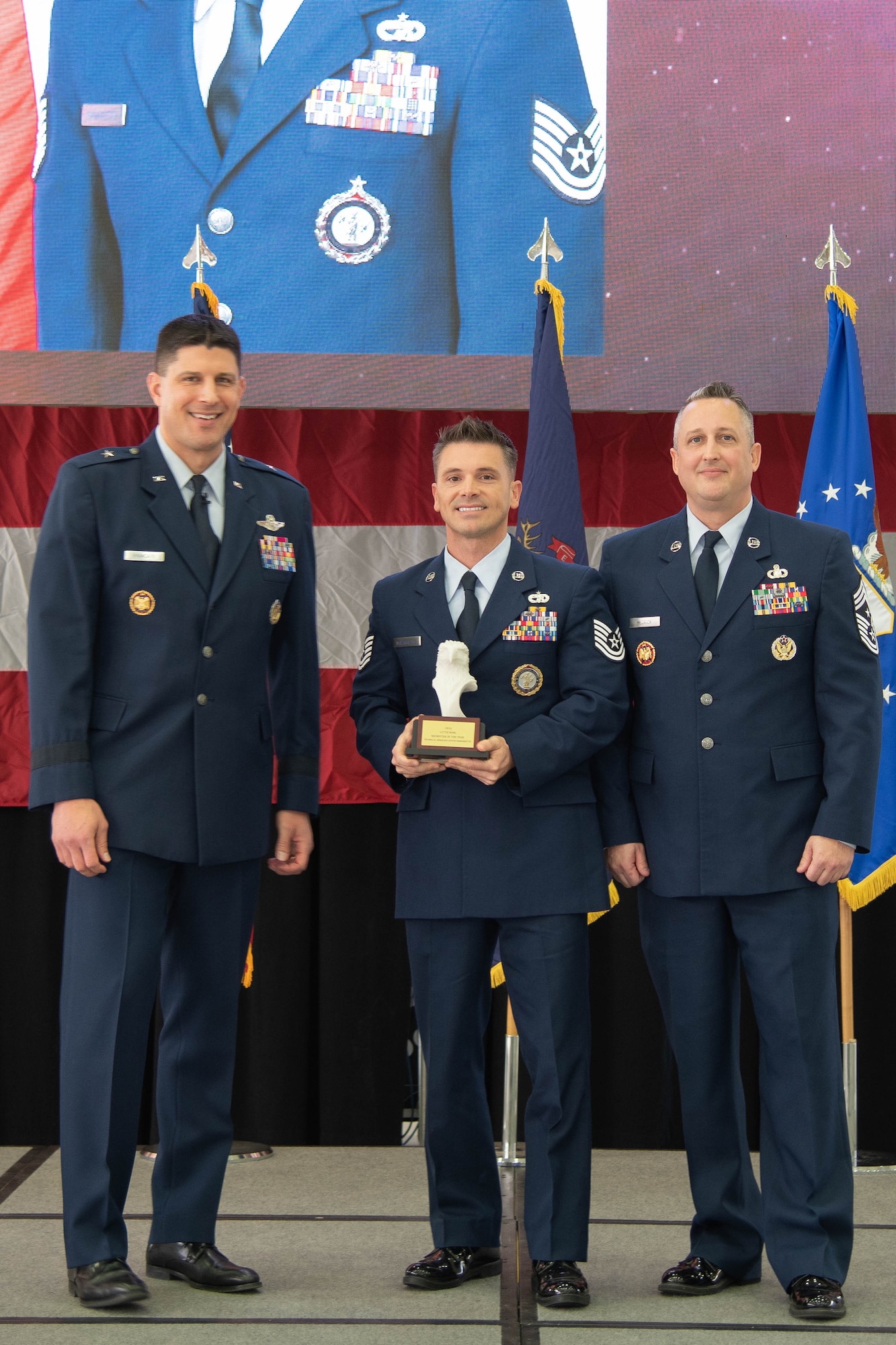 Air Force officer in dress blue uniform stands left of Air Force non-commissioned officer in dress blue uniform holding trophy, who stands left of Air Force senior enlisted leader in dress blue uniform .