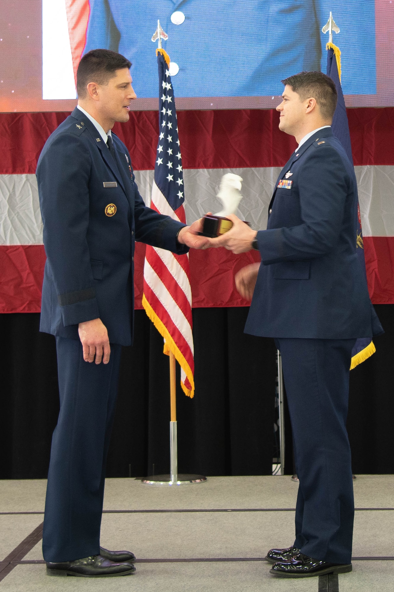 Air Force officer hands trophy to Air Force officer, both dressed in dress blue uniform.