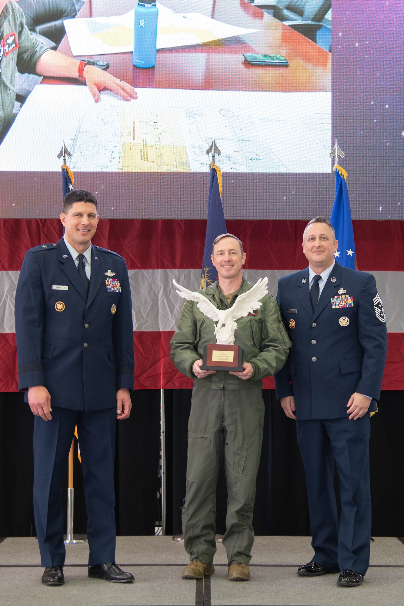 Air Force officer in dress blue uniform stands left of Air Force officer in flight suit holding large trophy, Air Force senior enlisted leader in dress blue uniform stands right.