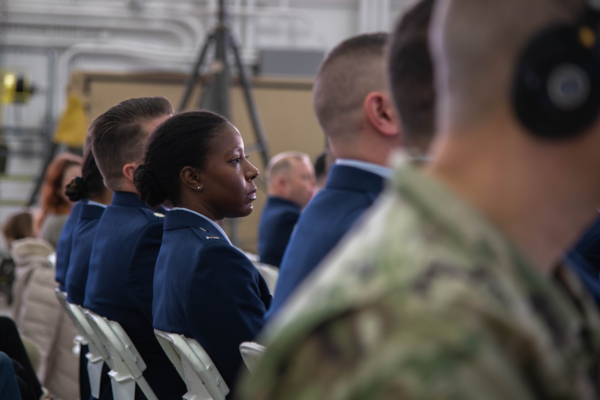 Female officer in dress blue uniform sits in audience, looking off to right of image.