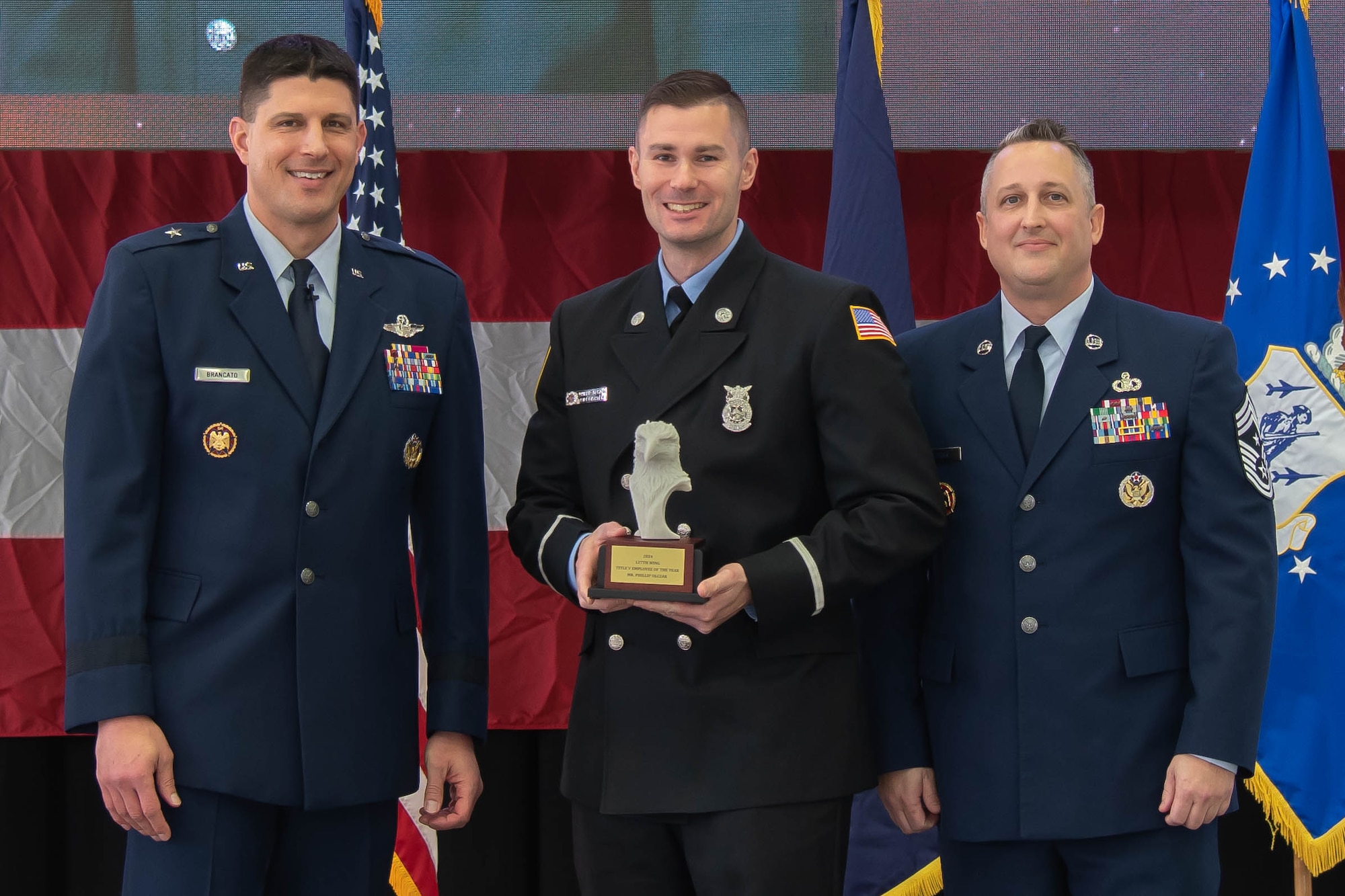 Air Force officer in dress blue uniform stands left of firefighter holding trophy, air force senior enlisted leader in dress blue uniform stands right.