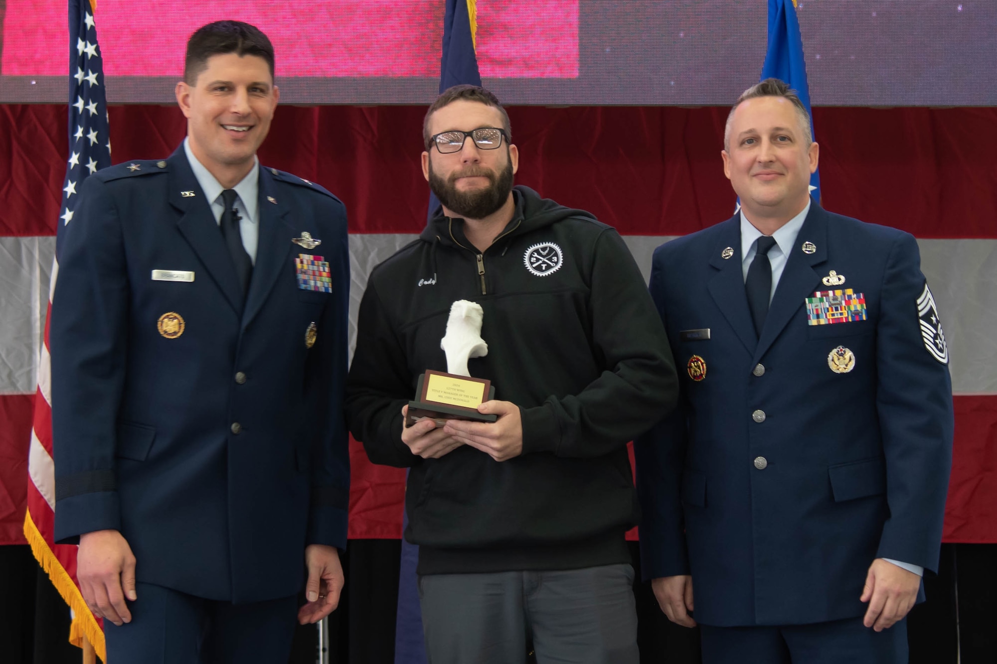 Air Force officer in dress blue uniform stands left of civilian man holding trophy, air force senior enlisted leader in dress blue uniform stands right.
